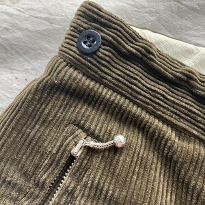 K&ouml;nnte beinhalten: Nahaufnahme von olivgr&uuml;nem Cordstoff mit Knopf, Rei&szlig;verschluss und Taschendetails. Der Stoff hat eine gerippte Textur. Der Rei&szlig;verschlussanh&auml;nger ist silberfarben mit einer kleinen Kugel am Ende. Der Knopf ist schwarz.