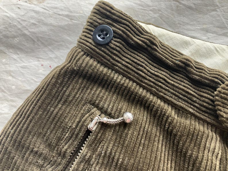 K&ouml;nnte beinhalten: Nahaufnahme von olivgr&uuml;nem Cordstoff mit Knopf, Rei&szlig;verschluss und Taschendetails. Der Stoff hat eine gerippte Textur. Der Rei&szlig;verschlussanh&auml;nger ist silberfarben mit einer kleinen Kugel am Ende. Der Knopf ist schwarz.