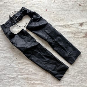 May include: Black leather chaps with a belt and buckle fastening. The chaps are displayed on a textured, off-white surface. The leather shows signs of wear, with visible stitching and a classic design.