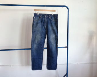 May include: Blue denim jeans hanging on a wooden hanger, suspended from a blue metal clothing rack. The jeans have a classic five-pocket design and a faded wash. The background is a plain white wall.