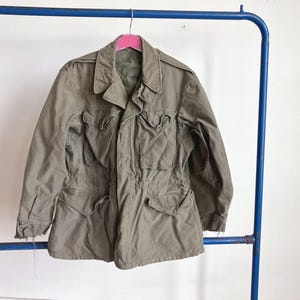 May include: An olive green military-style jacket with multiple front pockets and a collared neckline. The jacket is hanging on a pink hanger attached to a blue metal rack. The jacket has a worn appearance.