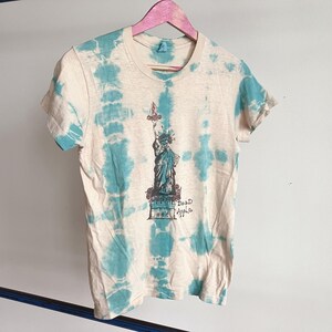 May include: Cream-colored t-shirt with a turquoise tie-dye pattern. The front features a graphic of the Statue of Liberty in blue and purple, with the text "Dead Apple" below. The shirt is hanging on a pink wooden hanger.