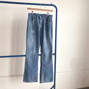 May include: Light blue corduroy pants hanging on a wooden hanger. The pants have a classic five-pocket design and a slight flare at the bottom. The corduroy fabric has a soft, textured appearance. The pants are displayed on a blue metal clothing rack.
