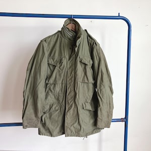 May include: An olive green military-style jacket with multiple front pockets and a high collar. The jacket is hanging on a blue metal clothing rack against a white wall. The jacket appears to be made of a durable, possibly water-resistant material.