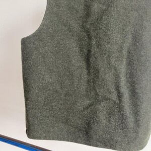 May include: A sleeveless vest made of a dark olive green felted material. The garment features a simple, solid-colored design with a rounded neckline and arm openings, showcasing a dense, textured fabric surface throughout the entire piece.