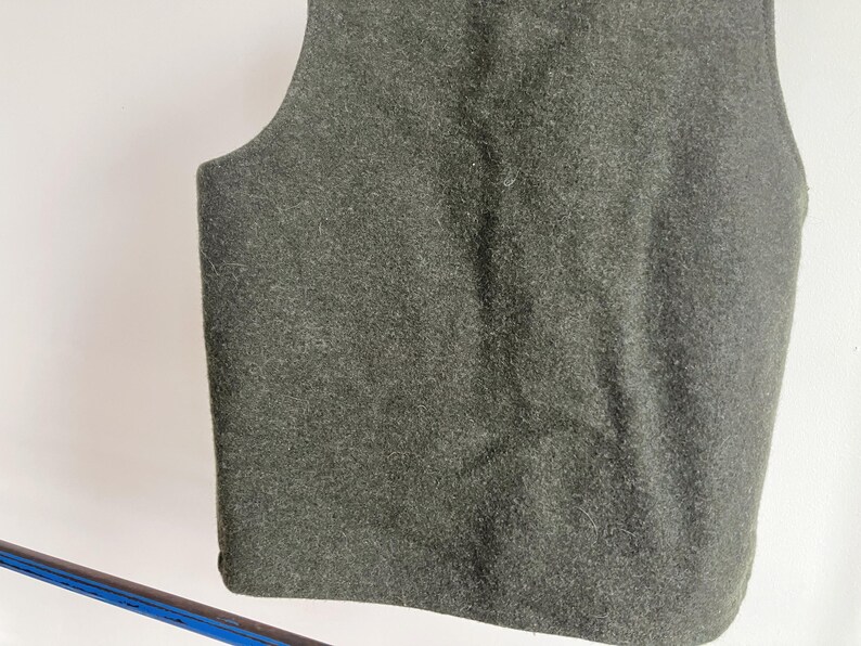 May include: A sleeveless vest made of a dark olive green felted material. The garment features a simple, solid-colored design with a rounded neckline and arm openings, showcasing a dense, textured fabric surface throughout the entire piece.