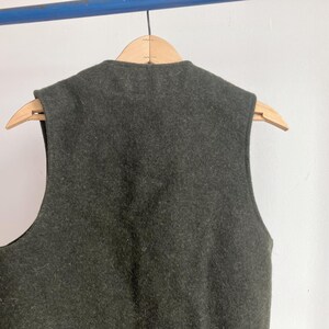 May include: A dark olive green sleeveless vest made of a thick, felted wool-like material. The garment features a high, rounded neckline and wide arm openings, hanging on a light-colored wooden hanger against a plain, light-colored wall.