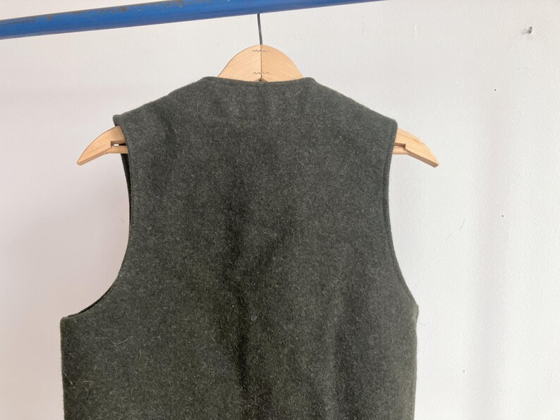 May include: A dark olive green sleeveless vest made of a thick, felted wool-like material. The garment features a high, rounded neckline and wide arm openings, hanging on a light-colored wooden hanger against a plain, light-colored wall.