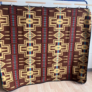 May include: A brown blanket with a geometric pattern in yellow, red, and turquoise. The design features repeating cross and diamond shapes. The blanket is displayed on a blue rack.