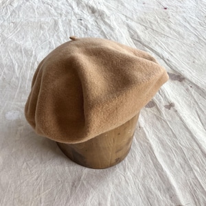 May include: A tan beret sits atop a wooden hat form. The beret is made of a soft, textured material and has a rounded crown. The hat form is cylindrical and made of light-colored wood. The background is a wrinkled, off-white fabric.