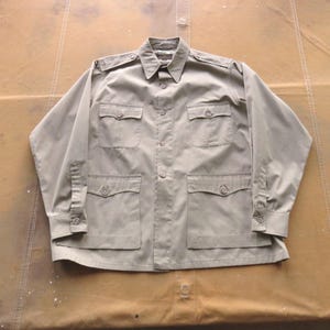 以下が含まれることがあります： ベージュの長袖シャツ。ボタン留めと複数のポケットが付いています。ゆったりとしたフィット感と襟があります。