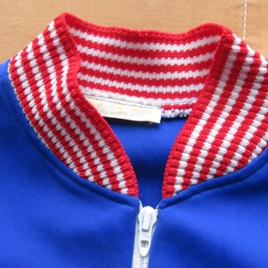 May include: A blue zippered jacket with a red and white striped collar. The collar is made of a knit fabric.