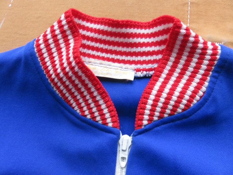 May include: A blue zippered jacket with a red and white striped collar. The collar is made of a knit fabric.
