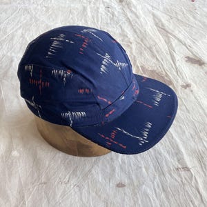 Könnte beinhalten: Eine marineblaue Kappe mit Schirm, verziert mit weißen und roten Pinselstrich-Designs. Die Stoffkappe liegt auf einem Holzständer. Das Muster wiederholt sich über die gesamte Oberfläche der Kappe. Ein modisches Accessoire.