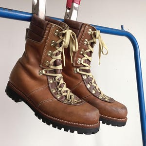 May include: A pair of tan and dark brown leather hiking boots featuring cream-colored laces, gold-tone metal eyelets, padded collars, and thick black rubber lug soles. The boots are suspended from a blue metal bar by silver metal clamps.