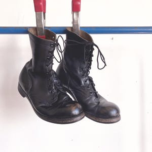 Puede incluir: Un par de botas de cuero negro con cordones, punteras redondeadas y cordones negros. Las botas tienen una altura hasta la pantorrilla, un tacón apilado y una construcción de vira visible. Están colgadas de una barra azul con dos pinzas metálicas.