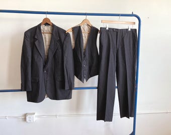 May include: A three-piece charcoal gray suit hanging on a blue rack. The suit includes a blazer, vest, and trousers. The blazer has two buttons and the vest has a button-down front. The trousers are straight-legged.