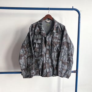 Könnte beinhalten: Eine Tarnjacke in Grau- und Brauntönen hängt an einem Holzbügel. Die Jacke hat mehrere Taschen und eine Knopfleiste vorne. Der Stoff weist ein blattartiges, abstraktes Design auf, das auf ein militärisches oder Outdoor-Thema hindeutet.