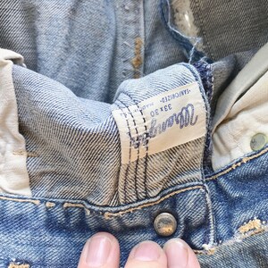 Peut inclure: Gros plan sur un jean Wrangler vintage. Le denim bleu clair montre des signes d'usure, avec une &eacute;tiquette visible affichant le nom de la marque et la taille 33x30. Le jean a une fermeture boutonn&eacute;e et une conception classique &agrave; cinq poches.