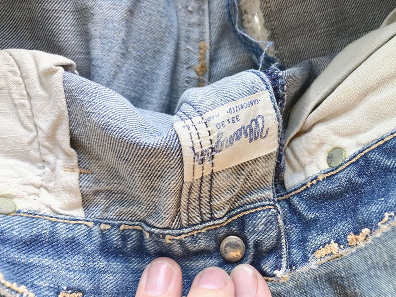 Peut inclure: Gros plan sur un jean Wrangler vintage. Le denim bleu clair montre des signes d'usure, avec une &eacute;tiquette visible affichant le nom de la marque et la taille 33x30. Le jean a une fermeture boutonn&eacute;e et une conception classique &agrave; cinq poches.