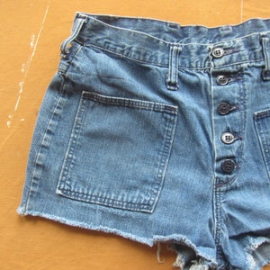 Op de afbeelding: Een blauwe denim short met een rafelige zoom en meerdere knoopsluitingen. De short heeft grote zakken aan de voorkant.