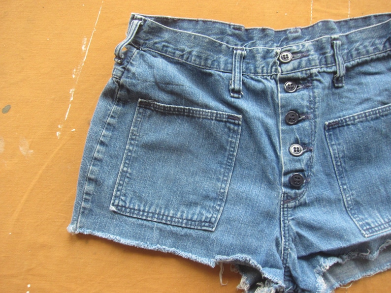 Op de afbeelding: Een blauwe denim short met een rafelige zoom en meerdere knoopsluitingen. De short heeft grote zakken aan de voorkant.