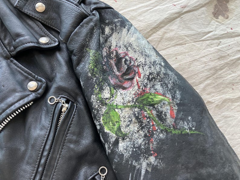 Op de afbeelding: Zwarte leren jas met een geschilderd rozenontwerp op de mouw. De roos is rood en groen, tegen een achtergrond van witte en grijze verfspatten. De jas heeft een ritssluiting en zilverkleurige hardware.