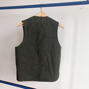 May include: A dark charcoal gray sleeveless vest made of a felted wool-like material. The garment features a simple round neckline and a straight hemline, hanging on a wooden hanger. The back of the vest is solid and plain with no visible closures.