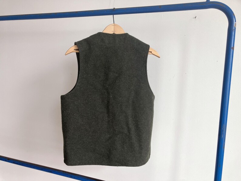 May include: A dark charcoal gray sleeveless vest made of a felted wool-like material. The garment features a simple round neckline and a straight hemline, hanging on a wooden hanger. The back of the vest is solid and plain with no visible closures.