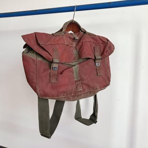 Może przedstawiać: Vintage plecak z czerwonego i oliwkowozielonego płótna z klapą i regulowanymi paskami. Torba zawieszona jest na metalowym wieszaku, prezentując zużytą fakturę i klasyczny design. Torba ma dwie klamry i uchwyt.