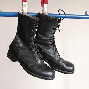 Op de afbeelding: Een paar zwarte leren gevechtslaarzen met hoge schacht, voorzien van zwarte veters door metalen oogjes, ronde neuzen en dikke zwarte rubberen zolen. De laarzen hangen aan twee rood-zilveren metalen klemmen aan een blauwe stang.