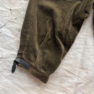 K&ouml;nnte beinhalten: Nahaufnahme einer olivgr&uuml;nen Cordhose. Die Hose hat eine gerippte Textur und ein dunkles Lederriemendetail am Kn&ouml;chel. Der Stoff ist gefaltet und zeigt die Textur- und Farbvariationen.
