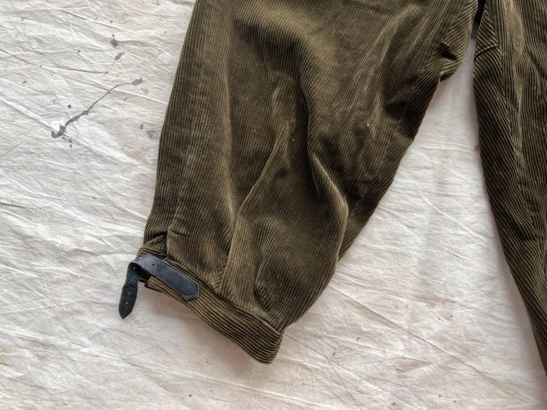 K&ouml;nnte beinhalten: Nahaufnahme einer olivgr&uuml;nen Cordhose. Die Hose hat eine gerippte Textur und ein dunkles Lederriemendetail am Kn&ouml;chel. Der Stoff ist gefaltet und zeigt die Textur- und Farbvariationen.