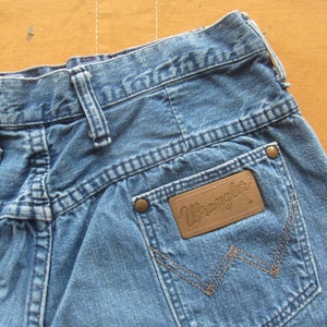 Op de afbeelding: Een paar blauwe denim jeans met een bruin leren patch met de tekst "Wrangler" op de achterzak.