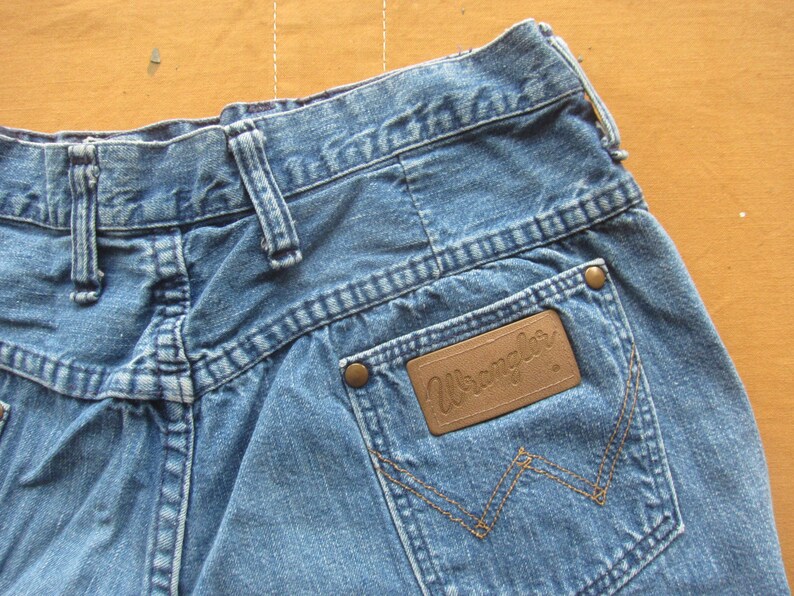 Op de afbeelding: Een paar blauwe denim jeans met een bruin leren patch met de tekst "Wrangler" op de achterzak.