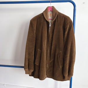 Puede incluir: Chaqueta de pana marrón con cierre de cremallera completo y dos bolsillos delanteros. La chaqueta tiene cuello y puños acanalados. Cuelga de una percha rosa contra una pared blanca.