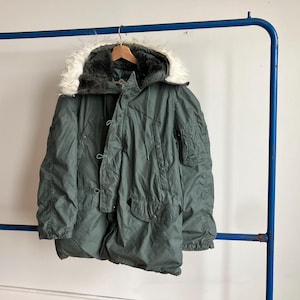 May include: An olive green vintage parka with a fur-lined hood hangs on a blue metal rack. The parka has multiple pockets, a button-up front, and a drawstring waist. The hood has a white fur trim.