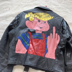 Op de afbeelding: Zwarte leren jas met een cartoonkarakterontwerp op de rug. Het personage heeft een roze huid, geel haar en draagt een blauw gestreept shirt en een rode overall. Het personage steekt zijn middelvinger op.