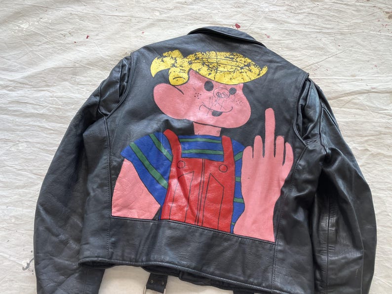 Op de afbeelding: Zwarte leren jas met een cartoonkarakterontwerp op de rug. Het personage heeft een roze huid, geel haar en draagt een blauw gestreept shirt en een rode overall. Het personage steekt zijn middelvinger op.