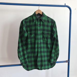 May include: A green and black plaid button-down shirt hangs on a wooden hanger. The long-sleeved shirt has two chest pockets and a classic collar. The shirt is displayed against a white background.