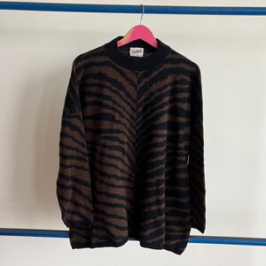 May include: A black and brown zebra-striped sweater with a mock neck. The sweater is hanging on a pink hanger against a white wall. The sweater has a tag on the neck.