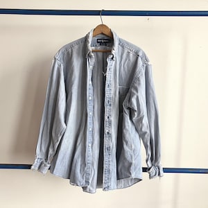 May include: Light blue denim button-down shirt with long sleeves. The shirt is hanging on a wooden hanger. The shirt has a button-down collar and a pocket on the left chest. The shirt is made of a light-wash denim material.