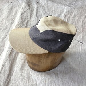 Könnte beinhalten: Beige und marineblaue Kappe mit gebogenem Schirm, auf einem Holzformteil. Die Kappe hat ein zweifarbiges Design, mit einem helleren beigen Stoff auf dem Schirm und den Vorderteilen und einem dunkleren marineblauen Stoff auf den Seitenteilen.