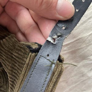 K&ouml;nnte beinhalten: Nahaufnahme eines besch&auml;digten, dunkelgrauen Lederriemens mit sichtbaren Abnutzungserscheinungen. Der Riemen ist an einem Abschnitt aus olivgr&uuml;nem Cordstoff befestigt. Das Leder weist Besch&auml;digungen auf, mit ausgefransten Kanten und L&ouml;chern.