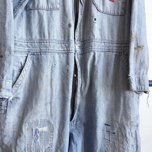 May include: Light blue denim jumpsuit with a full-length zipper, multiple pockets, and distressed details. The workwear-style garment features a red patch on the chest and frayed edges, showcasing a vintage aesthetic.