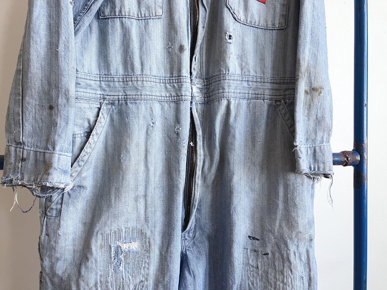 May include: Light blue denim jumpsuit with a full-length zipper, multiple pockets, and distressed details. The workwear-style garment features a red patch on the chest and frayed edges, showcasing a vintage aesthetic.