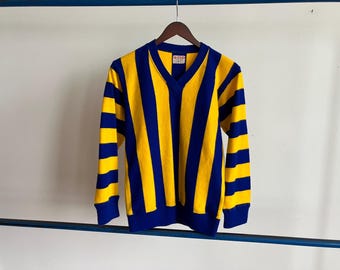 May include: A long-sleeved, V-neck shirt with vertical stripes in yellow and navy blue. The shirt is hanging on a wooden hanger against a white background. The brand name "SAND" is visible on the upper left.