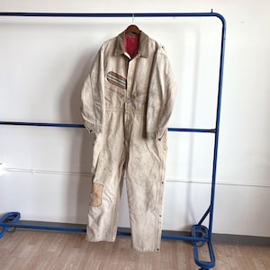 May include: A beige work jumpsuit with a red collar and a brown wooden hanger. The jumpsuit has a patch on the knee and a striped design on the chest pocket. It is hanging on a blue metal clothing rack.