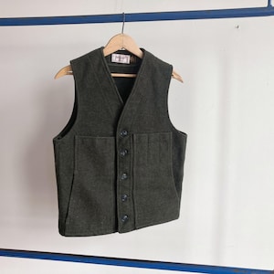 May include: A dark olive green wool vest featuring a V-neckline, a five-button front closure, and two large patch pockets on the front. The garment is displayed on a wooden hanger, showcasing a textured fabric finish and a visible interior label.