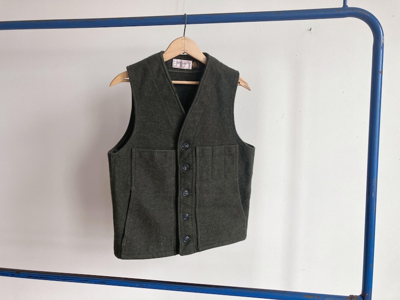 May include: A dark olive green wool vest featuring a V-neckline, a five-button front closure, and two large patch pockets on the front. The garment is displayed on a wooden hanger, showcasing a textured fabric finish and a visible interior label.